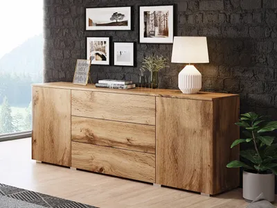 A wooden sideboard with drawers and doors is placed against a dark gray brick wall, with framed pictures and a lamp on top. A plant is visible to the right.