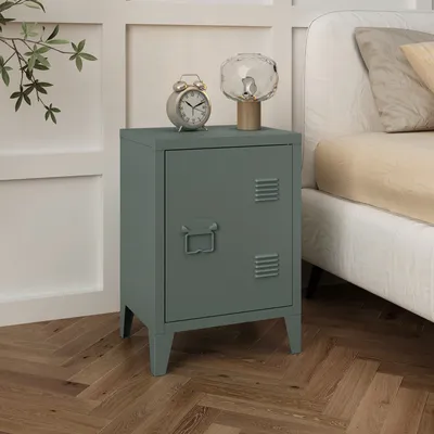 A dark gray metal nightstand with a locker-style door and ventilation slots, placed next to a bed. On top of the nightstand are a vintage-style alarm clock and a decorative glass lamp.