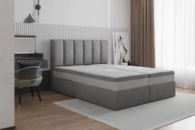 A bedroom scene featuring a large gray boxspring bed with a headboard, a desk with a chair, and curtains.