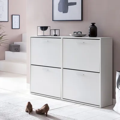A white shoe cabinet with four drawers is shown in a room with pink walls and a white floor. Two brown high-heeled shoes are placed on a rug in front of the cabinet.
