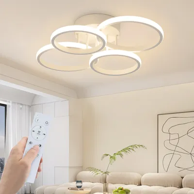 A hand holding a remote control is in the foreground, with a modern, multi-ringed ceiling light fixture illuminated above. The background shows a living room setting with a sofa and artwork.