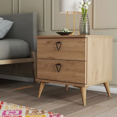 A bedside table with two drawers next to a bed, with a lamp and vase on top.