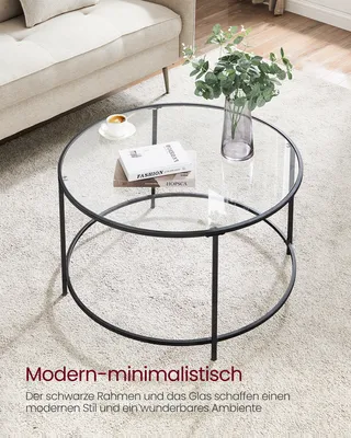 A round glass coffee table with a black metal frame is placed on a light-colored carpet next to a sofa. A vase with greenery and a cup of coffee are on the table.