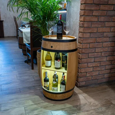A wooden barrel-shaped wine cabinet with several bottles of wine displayed inside, placed on a tiled floor next to a brick wall. A large plant is in the background.