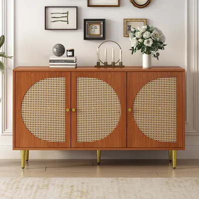 A wooden sideboard with rattan doors and gold legs is displayed against a white wall with framed pictures and a vase of flowers on top.