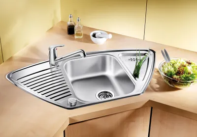 A modern stainless steel corner sink with a faucet, a drying rack, and a small compartment for herbs. A bowl of salad is placed on the countertop next to the sink.