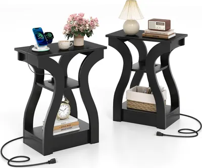 Two black side tables are shown against a white background. The tables have a modern, curved design and feature multiple shelves. The table on the left has a phone and smartwatch charging on its top surface, along with a cup and a vase of flowers. The tabl