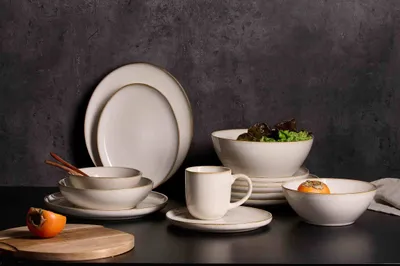 A collection of white stoneware tableware with gold trim, including plates, bowls, and a mug, arranged on a dark surface.