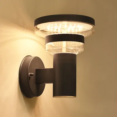 A modern black LED wall light with a clear glass shade is mounted on a light-colored wall.