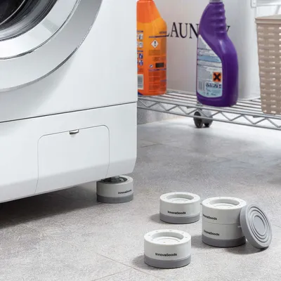A washing machine is shown with anti-vibration feet installed, and extra feet are placed on the floor nearby. Cleaning supplies are visible on a shelf in the background.