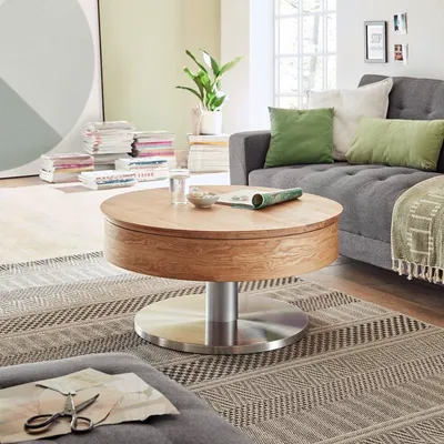 A modern living room with a round wooden coffee table in the foreground, a grey sofa with green cushions in the background, and stacks of books.