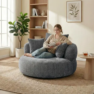 A woman is sitting on a large, round, gray sofa and reading a book in a well-lit living room.
