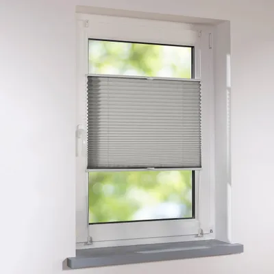 A close-up view of a gray pleated blind installed in a white window frame, with blurred green foliage visible through the window.