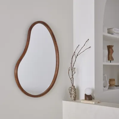 An organic-shaped mirror with a wooden frame hangs on a light gray wall next to a white shelf with decorative items.