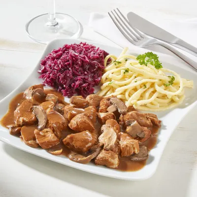 A plate of food containing goulash, red cabbage, and noodles, with cutlery and a wine glass in the background.