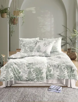 A bedroom scene with a bed dressed in green fern-patterned bedding, surrounded by hanging plants and natural light.