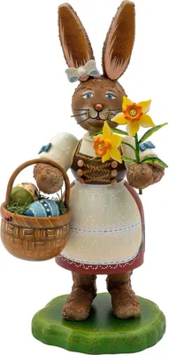 Ein fröhlicher Osterhase mit großen Ohren hält eine gelbe Blume in der rechten Hand und trägt in der linken einen Korb voller bunter Ostereier während er auf einem grünen Sockel steht und eine weiße Schürze mit blauen Details trägt