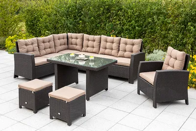 An outdoor patio set with a sectional sofa, armchair, table, and ottomans, set against a backdrop of lush green bushes and some yellow flowers.