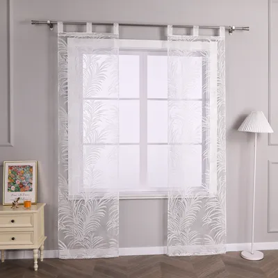 A window with white sheer curtains featuring a leaf pattern, a nightstand with a framed picture and a candle, and a floor lamp.