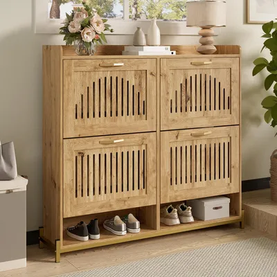 A wooden shoe cabinet with four doors and an open shelf at the bottom, displaying shoes and a decorative box. The cabinet is placed in an entryway or hallway setting with a plant and a lamp visible in the background.