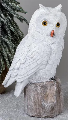A white owl figurine sits on a wooden stump, with a snowy evergreen tree in the background.