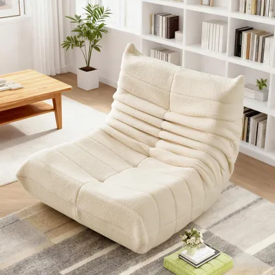 A beige, plush armchair is placed on a rug in a living room setting, with a wooden coffee table and bookshelves in the background.