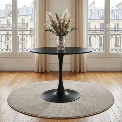 A round black dining table with a tulip base is placed on a beige rug in front of a large window with curtains.