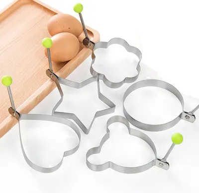 A set of five metal egg rings in different shapes, including a star, heart, round, flower, and bear, are arranged on a white surface next to two eggs on a wooden board.