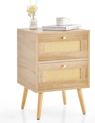 A light wood nightstand with two rattan-front drawers and gold knobs, shown against a white background. A small vase with a white flower and a book with a cup on top are placed on the surface.
