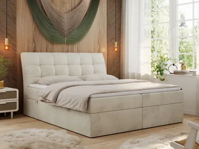 A bedroom scene featuring a beige boxspring bed with a tufted headboard, set against a wood-paneled wall with macrame wall art. A large window with sheer curtains is visible on the right, letting in natural light. A bedside table with drawers is on the lef