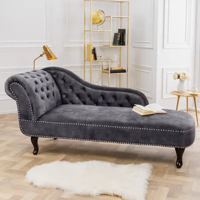 A gray velvet chaise lounge is placed on a wooden floor next to a white rug. A gold bookshelf with books and a globe is in the background.