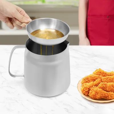A hand is pouring oil from a small metal bowl into a larger stainless steel pot. A plate of fried chicken is visible on the right side of the image.