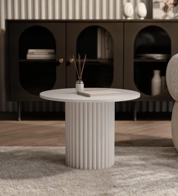 A round coffee table with a fluted base is placed on a rug in front of a dark cabinet.