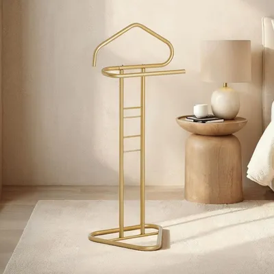 A gold valet stand is placed on a light-colored rug next to a wooden side table with a lamp and a book.