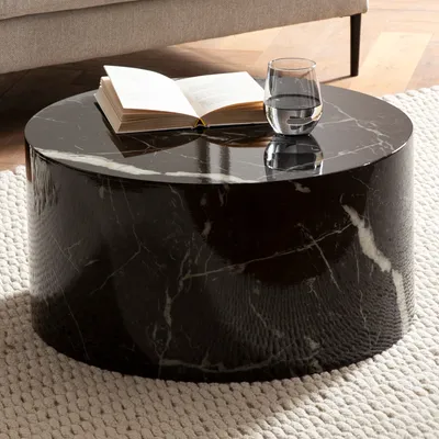 A round, black marble coffee table with a book and a glass of water on it, placed on a textured rug.