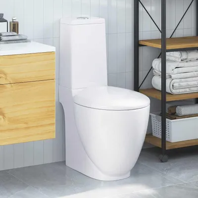 A white ceramic toilet in a modern bathroom setting, next to a wooden vanity and a shelving unit with towels.
