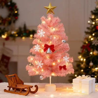 A pink artificial Christmas tree decorated with lights, ornaments, and a star topper, placed on a white surface with a wooden sled and a gift box nearby. Another Christmas tree is visible in the background.