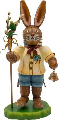 Ein fröhlicher Hase in traditioneller Kleidung hält einen Stab mit Blumen in der einen Hand und eine Glocke in der anderen während er auf einem grünen Sockel steht und freundlich lächelt