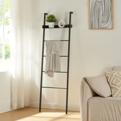 A black ladder shelf with a blanket draped over it, a small plant, and a vase on the top shelf, placed next to a window and a sofa in a living room.