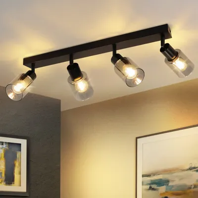 A black ceiling light fixture with four glass shades is mounted on a white ceiling. The light is on, casting a warm glow on the ceiling and walls. Two framed pictures are visible on the walls below the light.