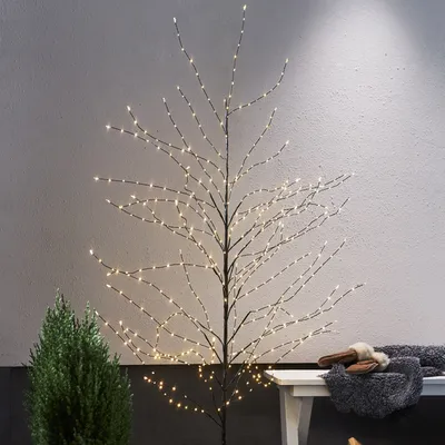 A decorative LED tree with warm white lights stands against a textured gray wall. A small green evergreen tree is to the left, and a white bench with a gray blanket and slippers is to the right.