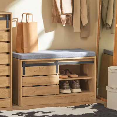 A shoe bench with storage is shown in a hallway or entryway setting, with clothes hanging in the background and a mirror reflecting the scene.