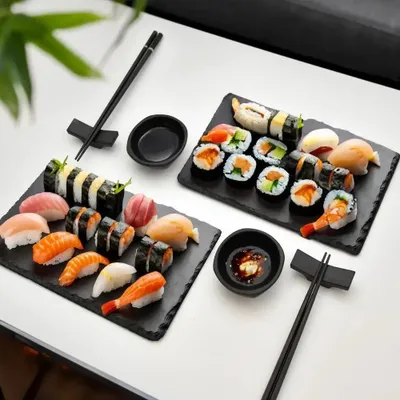 A sushi serving set with two black slate platters, sushi, chopsticks, and soy sauce dishes, arranged on a white surface.