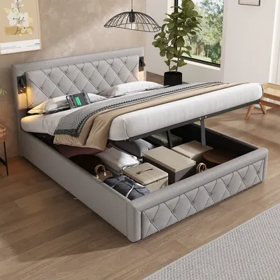 A bedroom scene featuring a hydraulic storage bed with its storage compartment open, revealing stored items. The bed is made up with bedding and pillows, and a plant is visible in the background.
