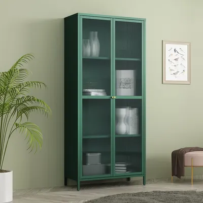 A green glass-door cabinet filled with decorative items, placed in a room with a plant and a framed picture on the wall.