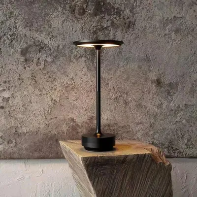 A black LED table lamp is placed on a wooden block against a textured gray wall.