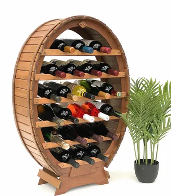 A wooden wine rack shaped like a barrel, filled with various bottles of wine, stands against a white background. A potted plant with green fronds is placed to the right of the wine rack.