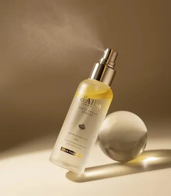 A bottle of d'Alba First Spray Serum is shown with mist spraying from the nozzle, next to a clear sphere on a gradient background.