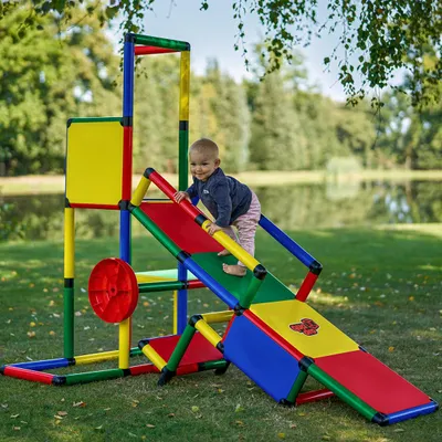 A baby is playing on a colorful playground structure in a grassy outdoor area with a lake in the background.
