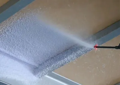 A spray nozzle is spraying a white foam onto a surface, likely for insulation.